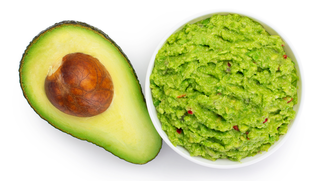 Bowl of fresh guacamole next to half of an avocado with the pit.