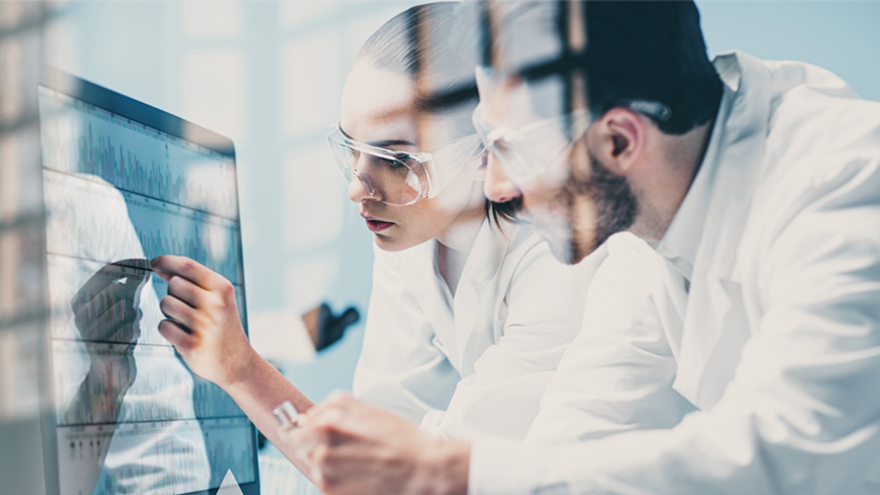 Two pharmaceutical scientists wearing white coats and goggles performing statistical process control on a computer.
