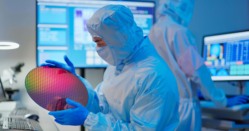 Semiconductor technician wearing full cleanroom protective gear and blue gloves inspecting a reflective silicon wafer, with digital monitoring screens and another technician working in the background.